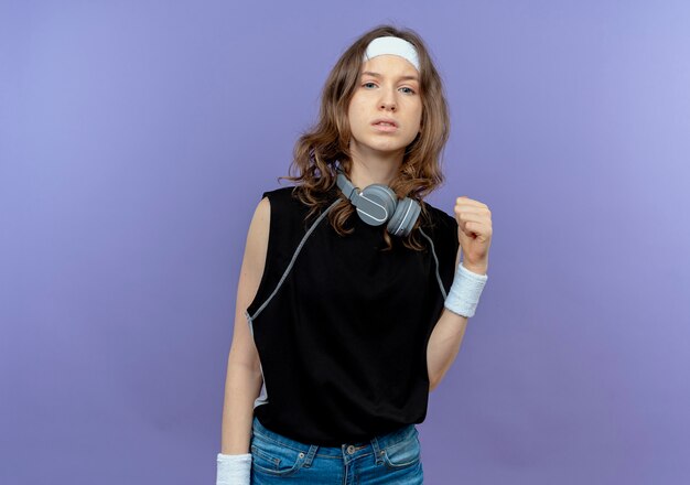 Young fitness girl in black sportswear with headband looking confident clenching fist standing over blue wall