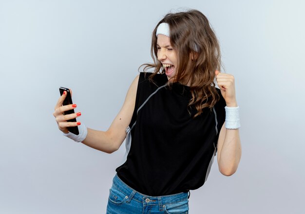 Молодая фитнес-девушка в черной спортивной одежде с повязкой на голову, смотрящая на экран своего смартфона, счастливая и взволнованная сжимая кулак, стоящая над белой стеной