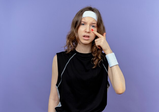 Young fitness girl in black sportswear with headband  pointing with finger to her eye standing over blue wall