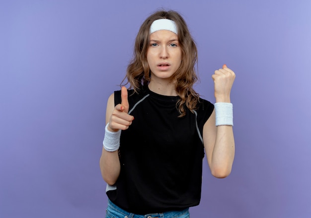 Young fitness girl in black sportswear with headband pointing with index finger clenching fist looking with serious face standing over blue wall