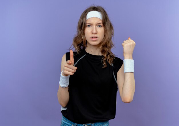 Young fitness girl in black sportswear with headband pointing with index finger clenching fist looking with serious face standing over blue wall