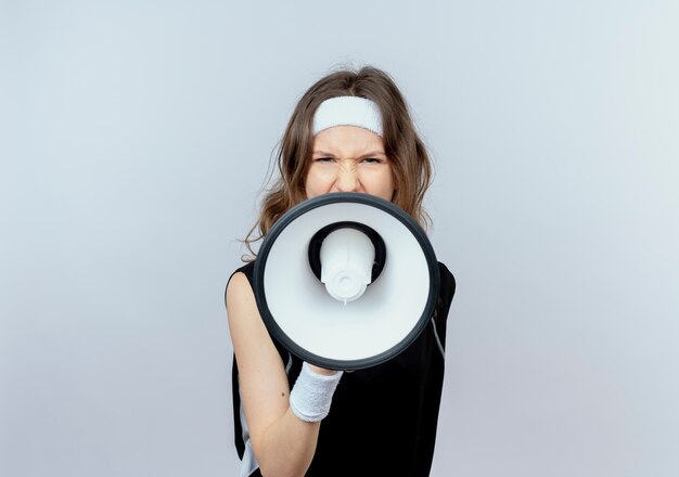 Young fitness girl in black sportswear with headband shouting to megaphone with aggressive expression standing over white wall