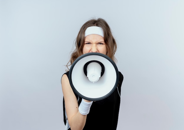 Молодая фитнес-девушка в черной спортивной одежде с повязкой на голове кричит в мегафон с агрессивным выражением лица, стоя над белой стеной