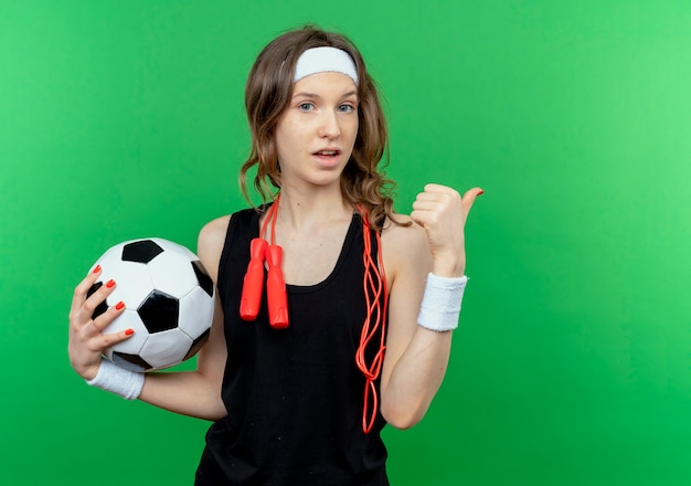 Foto gratuita giovane ragazza di forma fisica in abiti sportivi neri con fascia e corda per saltare intorno al collo tenendo il pallone da calcio confuso che punta indietro in piedi sopra la parete verde