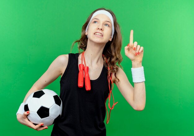 Молодая фитнес-девушка в черной спортивной одежде с повязкой на голову и скакалкой на шее держит футбольный мяч, глядя вверх, показывая указательный палец, улыбаясь над зеленым