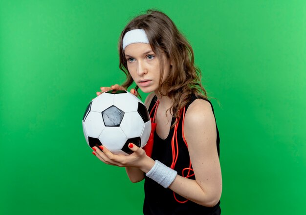 Young fitness girl in black sportswear with headband and skipping rope around neck holding soccer ball looking with serious expression standing over green wall