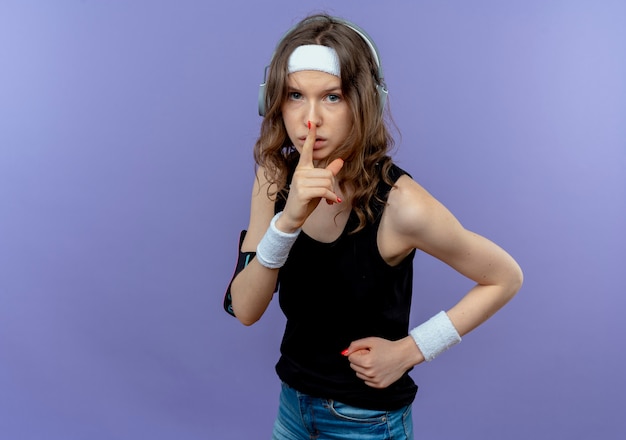 Молодая фитнес-девушка в черной спортивной одежде с повязкой на голову и повязкой для смартфона, делая жест молчания с пальцем на губах, стоя над синей стеной