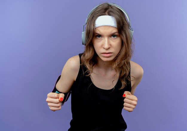 Young fitness girl in black sportswear with headband and smartphone armband with clenched fists strained and confident standing over blue wall