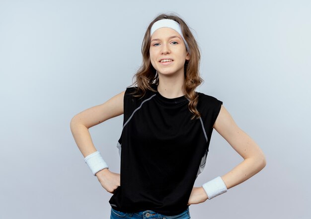 Young fitness girl in black sportswear with headband  smiling confident with arms at hip standing over white wall