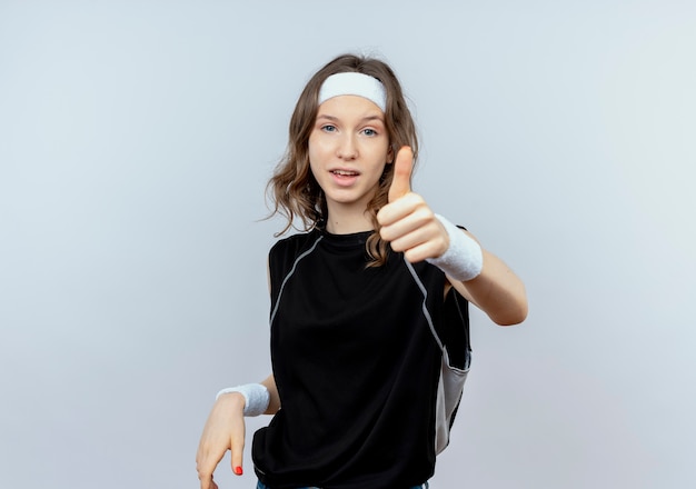 Young fitness girl in black sportswear with headband  smiling showing thumbs up standing over white wall