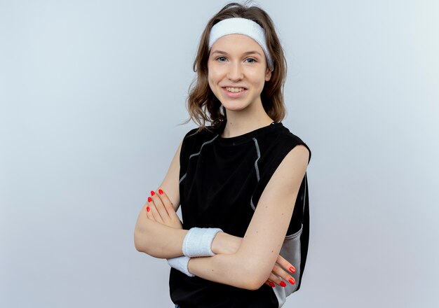 Young fitness girl in black sportswear with headband  smiling with crossed hands standing over white wall