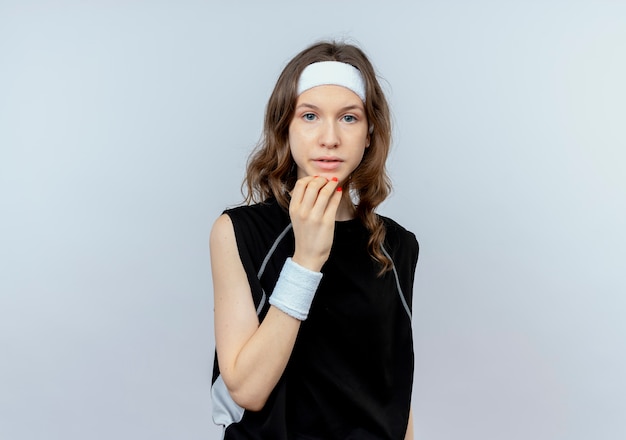 Young fitness girl in black sportswear with headband  stressed and nervous standing over white wall