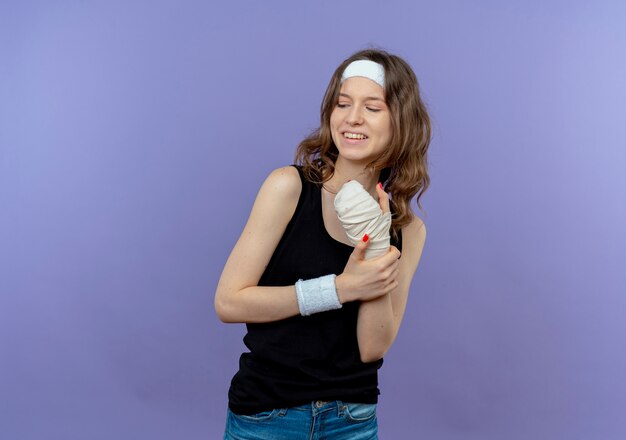 Молодая фитнес-девушка в черной спортивной одежде с повязкой на голове, касаясь ее перевязанного запястья, выглядит смущенной, стоя у синей стены