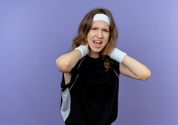 Young fitness girl in black sportswear with headband touching her neck looking displeased having pain standing over blue wall