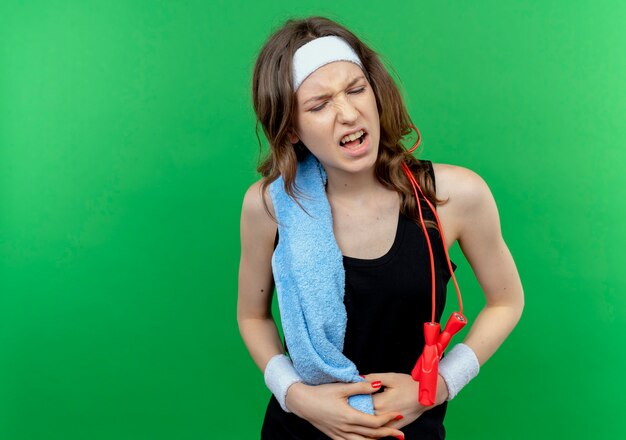 Young fitness girl in black sportswear with headband and towel on shoulder looking unwell touching her belly feeling pain standing over green wall