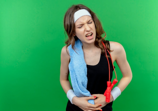 Young fitness girl in black sportswear with headband and towel on shoulder looking unwell touching her belly feeling pain standing over green wall