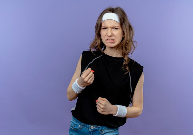 Young fitness girl in black sportswear with headband  with angry face clenching fists standing over blue wall