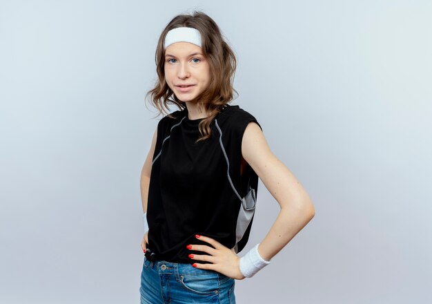 흰 벽 위에 서있는 자신감이 심각한 표정으로 머리띠와 검은 운동복에 젊은 피트니스 소녀