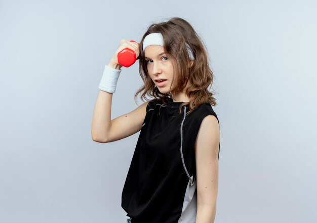 Молодая фитнес-девушка в черной спортивной одежде с повязкой на голову, тренируясь с гантелями, уверенно смотрит, стоя над белой стеной