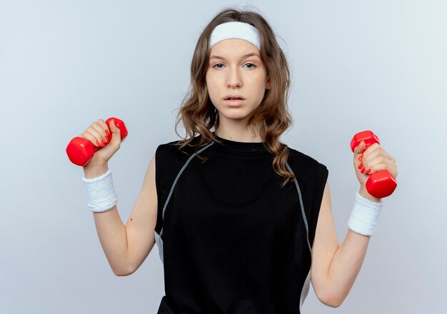 Молодая фитнес-девушка в черной спортивной одежде с повязкой на голову, тренирующаяся с гантелями, уверенно выглядит, стоя над белой стеной