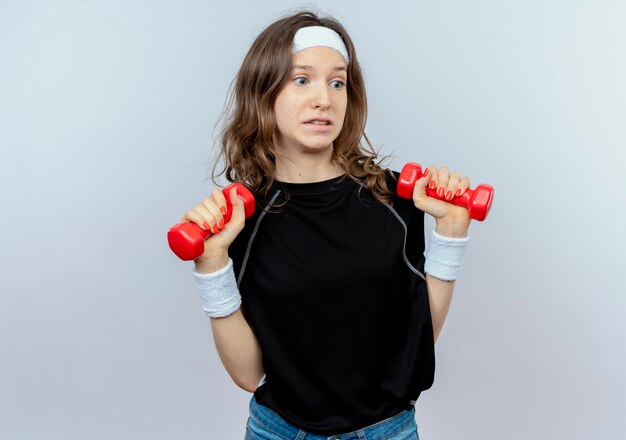 Молодая фитнес-девушка в черной спортивной одежде с повязкой на голову, тренирующаяся с гантелями, выглядит смущенной, стоя над белой стеной