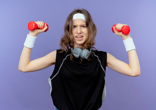 Young fitness girl in black sportswear with headband working out with dumbbells looking strained over blue