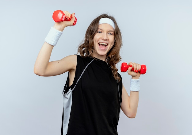 Young fitness girl in black sportswear with headband working out with dumbbells  smiling cheerfully standing over white wall