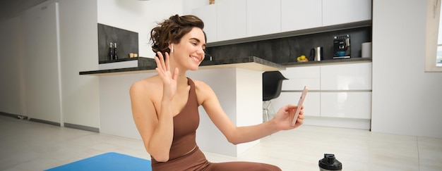 Free photo young fitness girl at home sport instructor in tracksuit waves hand at smartphone sits on workout