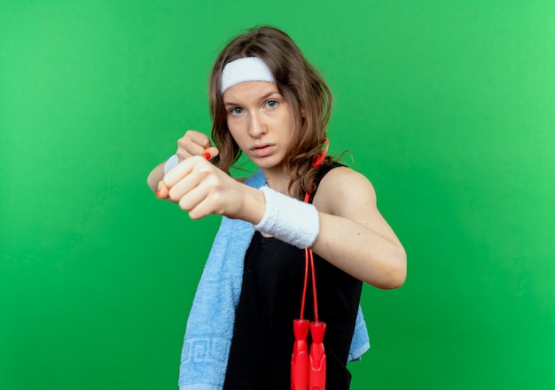 Бесплатное фото Молодая фитнес-девушка в черной спортивной одежде с повязкой на голову и полотенцем на плече со сжатыми кулаками, как боксер, стоящий над зеленой стеной