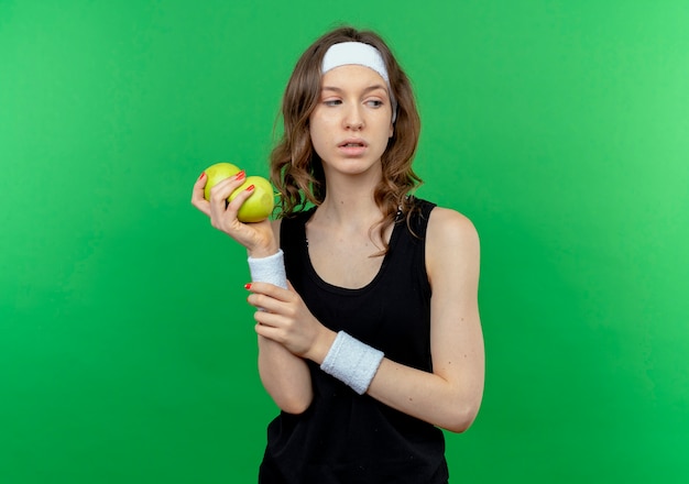 Бесплатное фото Молодая фитнес-девушка в черной спортивной одежде с повязкой на голове держит два зеленых яблока, скептически глядя в сторону, стоя у зеленой стены