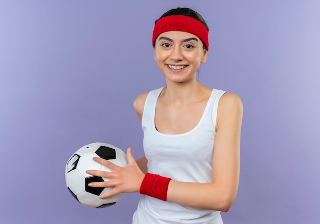 Бесплатное фото Молодая фитнес-девушка в спортивной одежде с повязкой на голову, держащая футбольный мяч, уверенно улыбается, счастлива и позитивно стоит над фиолетовой стеной