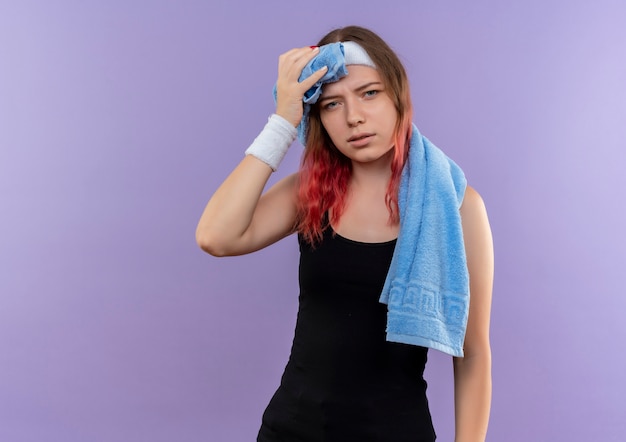 Бесплатное фото Молодая фитнес-девушка в спортивной одежде с полотенцем на шее выглядит усталой и измученной, стоя у фиолетовой стены