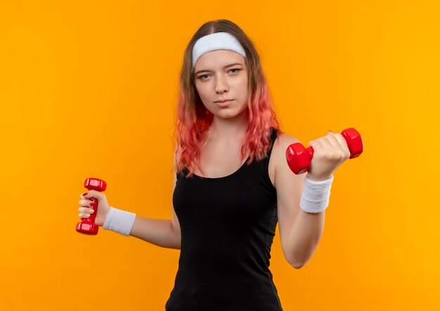 Young fitness girl in sportswear using dumbbells doing exercises looking confident standing over orange wall