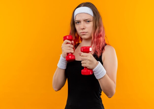 Young fitness girl in sportswear using dumbbells doing exercises looking confident standing over orange wall