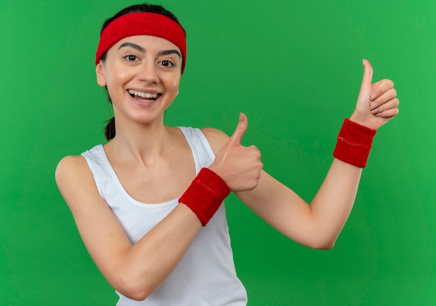 Young fitness girl in sportswear with headband  happy and positive smiling cheerfully showing thumbs up standing over green wall