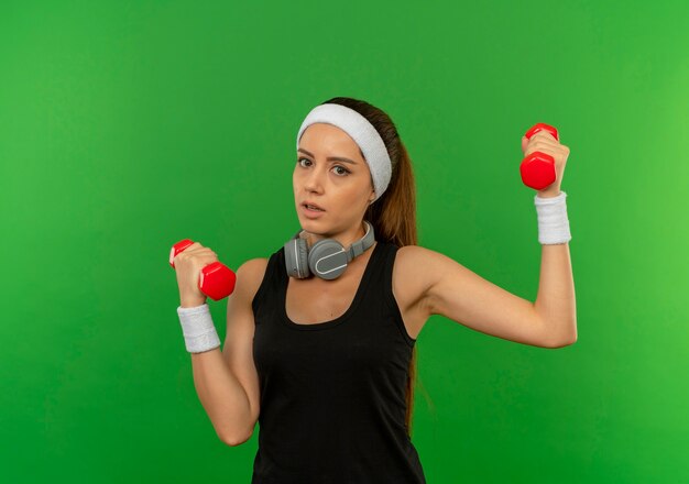 Young fitness girl in sportswear with headband holding two dumbbells doing exercises looking confused standing over green wall