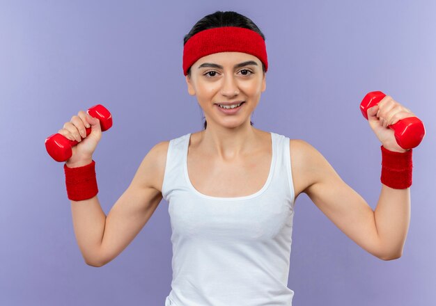 Young fitness girl in sportswear with headband holding two dumbbells in raised hands smiling cheerfully doing exercises standing over purple wall