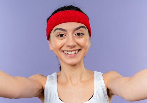 Молодая фитнес-девушка в спортивной одежде с повязкой на голову, весело улыбаясь, делая приветственный жест руками, стоящими над фиолетовой стеной