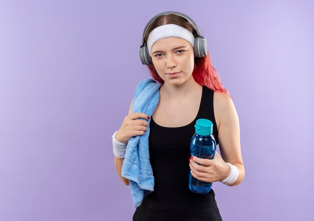 紫色の壁の上に立っている水のボトルを保持している彼女の首にヘッドフォンとタオルでスポーツウェアの若いフィットネスの女の子