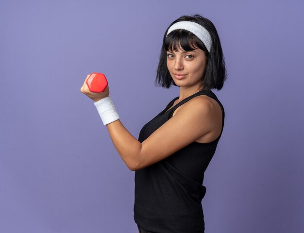 Young fitness girl wearing headband holding dumbbell doing exercises looking confident smiling standing over blue background