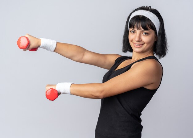Young fitness girl wearing headband holding dumbbells doing exercises looking confident smiling standing over white