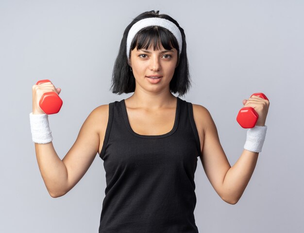 Young fitness girl wearing headband holding dumbbells doing exercises looking confident standing over white background