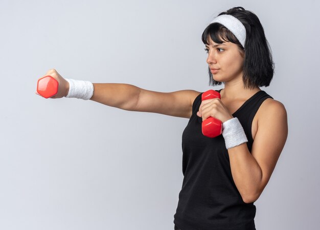 Young fitness girl wearing headband holding dumbbells doing exercises looking confident standing over white background