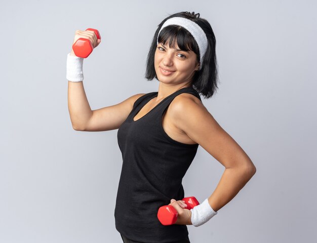 Young fitness girl wearing headband holding dumbbells doing exercises looking confident standing over white