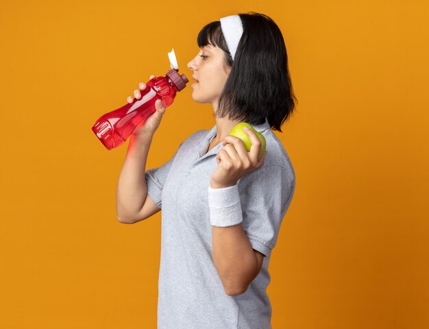 オレンジ色の背景の上に立っているボトルから新鮮なリンゴの飲料水を保持しているヘッドバンドを身に着けている若いフィットネスの女の子