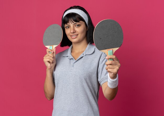 Young fitness girl wearing headband holding rackets for table tennis looking at camera with smile on face standing over pink background