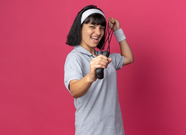 Young fitness girl wearing headband holding skipping rope looking at camera happy and cheerful smiling standing over pink background