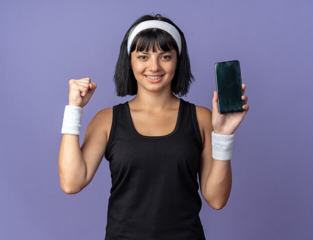 Young fitness girl wearing headband holding smartphone clenching fist happy and excited looking at camera 