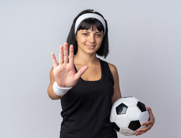 Young fitness girl wearing headband holding soccer ball looking at camera smiling doing stop gesture with hand standing over white