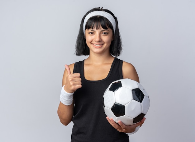 サッカーボールを保持しているヘッドバンドを身に着けている若いフィットネスの女の子は、白の上に立って買い物をしている親指を笑顔のカメラを見て